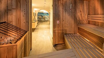 Ein gemütlicher Saunabereich aus Holz mit Bänken und einem Eingang zu einem weiteren Raum. Sanfte Beleuchtung schafft eine entspannte Atmosphäre.