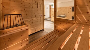 Ein gemütlicher Saunabereich aus Holz mit einer warmen und einladenden Atmosphäre. Im Hintergrund sind weitere Holz-Elemente und Duschen zu sehen.