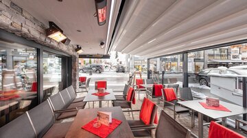 Ein modernes Restaurant mit Tischen und Stühlen im Innenbereich. Rote Tischdecken und Heizstrahler schaffen eine einladende Atmosphäre.