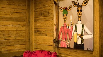 Ein künstlerisches Bild von zwei stilisierten Rehen, die in Tracht gekleidet sind. Die Wand besteht aus Holzpaneelen, und im Vordergrund steht ein rotes Sofa.