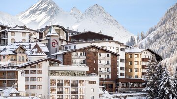Ein malerisches Winterdorf mit schneebedeckten Bergen im Hintergrund. Modern gestaltete Gebäude und ein klarer, blauer Himmel schaffen eine gemütliche Atmosphäre.