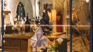 Ein schön gedeckter Tisch mit Blumen und dekorativen Elementen ist durch ein Fenster sichtbar. Im Hintergrund sind warme Lichtquellen und rustikale Möbel zu sehen.