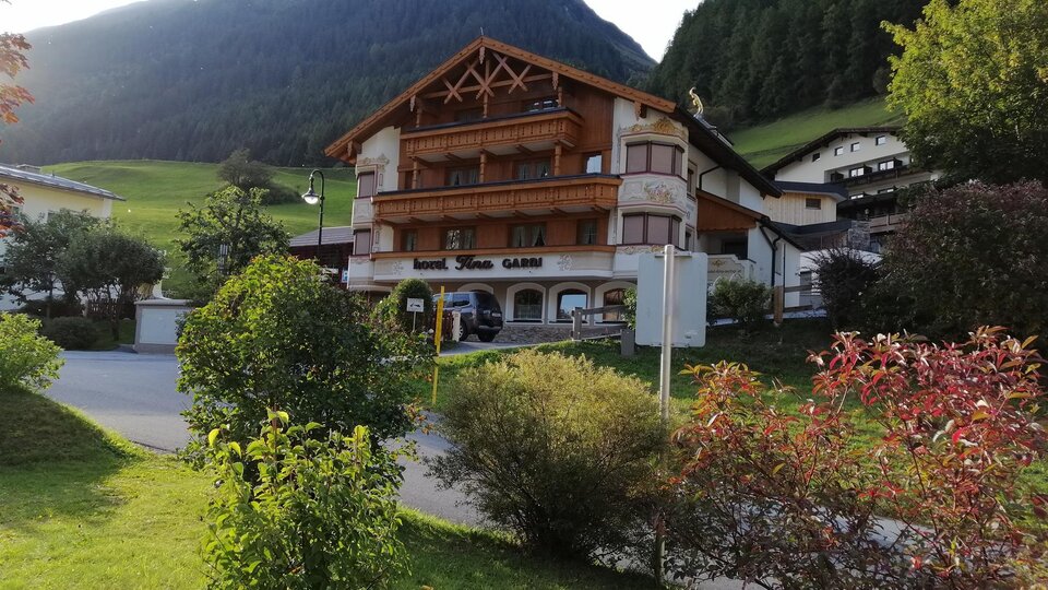 Ein charmantes Holzhaus inmitten von grünen Wiesen und Bergen. Vor dem Gebäude wachsen blühende Sträucher und Bäume.