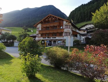 Ein charmantes Holzhaus inmitten von grünen Wiesen und Bergen. Vor dem Gebäude wachsen blühende Sträucher und Bäume.
