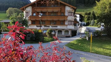 Ein rustikales Hotel mit Holzverkleidung, umgeben von grünen Bergen. Sträucher mit roten Blättern im Vordergrund.