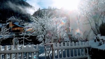 Ein verschneiter Garten mit einem Holzzaun und frostigen Bäumen. Die Sonne scheint hell über die Berge.
