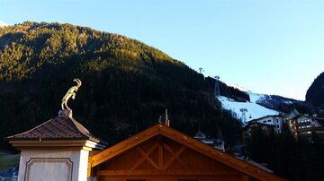 Eine malerische Berglandschaft mit schneebedeckten Hängen und einer Seilbahn. Im Vordergrund ist ein traditionelles Gebäude mit einer dekorativen Statue zu sehen.