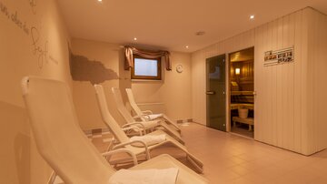 Ein entspannender Wellnessbereich mit Liegen und einer Sauna. Die Wände sind hell und die Beleuchtung ist sanft.