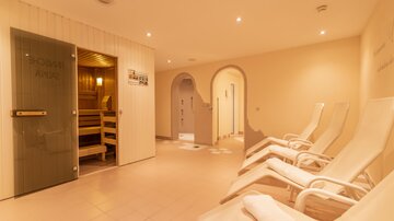Ein moderner Wellnessbereich mit Liegen und einer Sauna. Die sanften Farben und das dezente Licht schaffen eine entspannende Atmosphäre.