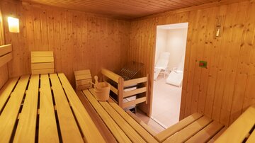 Eine gemütliche Sauna aus Holz mit Bänken und einem Eimer für Wasser. Der Raum strahlt Wärme und Entspannung aus.