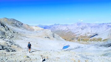 Ein Wanderer erkundet eine felsige Berglandschaft mit einem klaren blauen Himmel. Im Hintergrund sind majestätische Berge und ein kleiner See zu sehen.