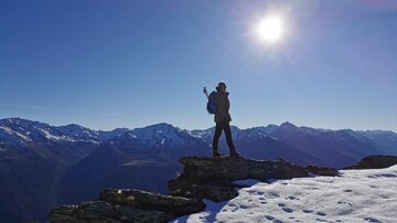Eine Person steht auf einem Felsen mit Blick auf schneebedeckte Berge. Die Sonne scheint hell am blauen Himmel.