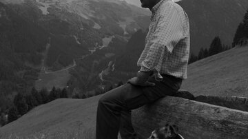 Ein Mann sitzt auf einem Baumstamm und blickt auf eine bergige Landschaft. Neben ihm liegt ein Hund, während die Wolken den Himmel bedecken.
