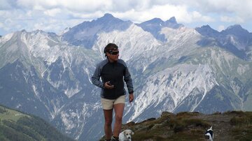 Eine Person wandert in den Bergen mit zwei Hunden. Im Hintergrund sind majestätische Berggipfel und eine hügelige Landschaft zu sehen.