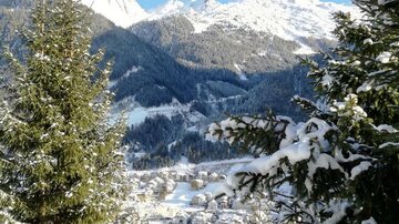 Eine winterliche Landschaft mit schneebedeckten Bergen und Tannenbäumen. Im Hintergrund ist ein Dorf zu sehen, umgeben von der schönen Natur.
