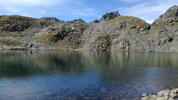 Ein ruhiger Bersee umgeben von felsigen Bergen. Der Himmel ist blau mit einigen Wolken.