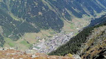 Ein Blick auf die malerische Tallandschaft mit einem kleinen Dorf. Umgeben von grünen Hügeln und hohen Bergen.