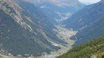 Ein malerisches Tal umgeben von hohen Bergen. Im Hintergrund sind ein kleines Dorf und klare, grüne Wiesen zu sehen.