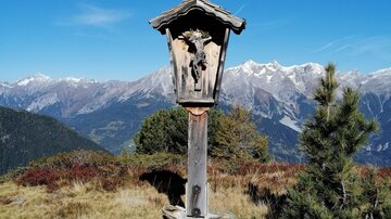 Ein Holzschild steht in der Natur mit schneebedeckten Bergen im Hintergrund. Die Landschaft ist grün und der Himmel ist klar und blau.