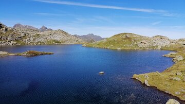 Ein ruhiger Bergsee umgeben von grünen Wiesen und felsigen Hügeln. Der Himmel ist klar und blau, was eine friedliche Stimmung schafft.