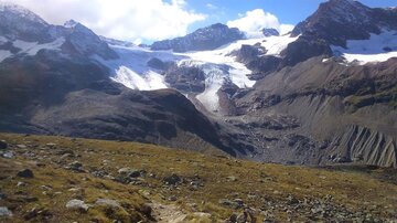 Ein beeindruckendes Bergpanorama mit schneebedeckten Gipfeln und glazialen Formationen. Im Vordergrund ist eine grüne Wiese mit Steinen sichtbar.