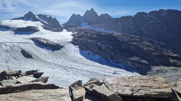 Eine beeindruckende Berglandschaft mit schneebedeckten Gipfeln und klaren blauen Himmel. Im Vordergrund sind große Felsen sichtbar.