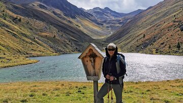 Eine Person steht neben einem Wegweiser in einer Berglandschaft. Im Hintergrund ist ein klarer See und majestätische Berge zu sehen.