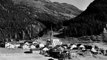 Ein malerisches Bergdorf mit traditionellen Häusern und einer Kirche im Vordergrund. Die umliegenden Berge sind mit Bäumen bewachsen und prägen die idyllische Landschaft.