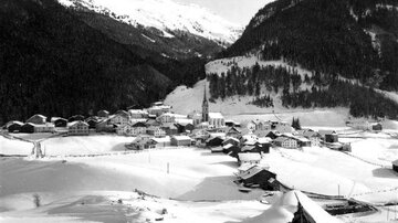 Ein malerisches Dorf im Schnee mit traditionellen Häusern und einer Kirche. Die umgebende Landschaft ist von Bergen und Wäldern geprägt.