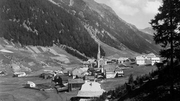 Ein malerisches Dorf in den Bergen, umgeben von hohen Tannen und weiten Feldern. Die Kirche mit dem spitzen Turm dominiert die Landschaft.