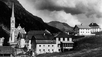Eine historische Schwarz-Weiß-Aufnahme eines malerischen Dorfes in den Bergen. Die Kirche und traditionelle Gebäude sind sichtbar, umgeben von felsigen Hügeln und dramatischem Himmel.