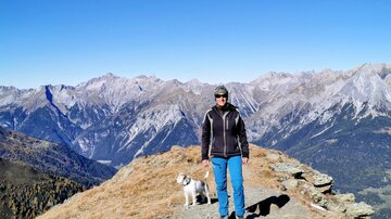 Eine Person steht auf einem Aussichtspunkt in den Bergen, neben einem Hund. Der Himmel ist klar und die Berge erstrecken sich in der Ferne.