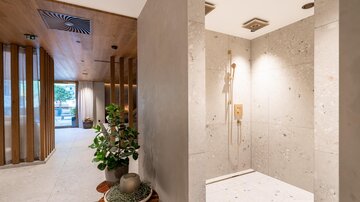 Ein modernes Badezimmer mit einer Dusche aus Stein und eleganter Ausstattung. Im Hintergrund sind Pflanzen und ein hell erleuchteter Raum sichtbar.