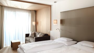 Ein modernes Hotelzimmer mit zwei Betten und einer gemütlichen Sitzecke. Große Fenster sorgen für viel Tageslicht.