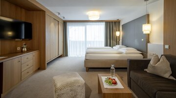 Ein modernes Hotelzimmer mit zwei Einzelbetten, einem Fernseher und einem bequemen Sofa. Große Fenster sorgen für viel natürliches Licht.