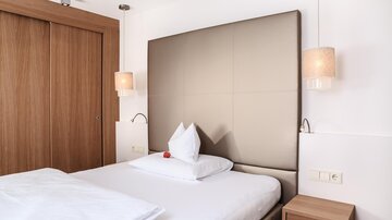 Ein modernes Schlafzimmer mit einem großen Bett und gemütlicher Beleuchtung. Die Wände sind in neutralen Farben gehalten und es gibt eine Holzschranktür.