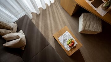 Ein gemütlicher Wohnbereich mit einem Sofa und einem kleinen Tisch. Auf dem Tisch liegt eine Obstschale und Sonnenlicht scheint durch die Vorhänge.