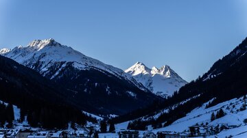 Eine ruhige Winterlandschaft mit schneebedeckten Bergen und einem klaren blauen Himmel. Im Vordergrund sind kleine Häuser in einem Tal sichtbar.