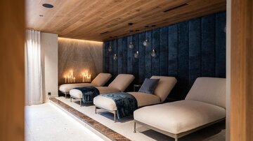 Ein moderner Entspannungsraum mit bequemen Liegen und sanfter Beleuchtung. Die Wände sind geheimnisvoll in dunklem Blau gehalten, was eine ruhige Atmosphäre schafft.