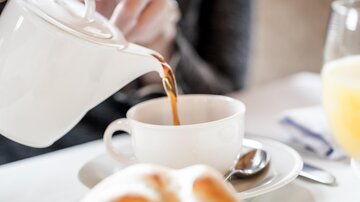 Eine Hand gießt frisch gebrühten Tee in eine weiße Tasse. Im Vordergrund liegt ein Brötchen auf einem Teller.