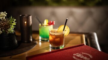 Zwei Cocktails auf einem Holztisch in einer gemütlichen Umgebung. Im Vordergrund ein Drink mit einer Orange und im Hintergrund ein grüner Cocktail.