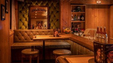 Eine gemütliche Bar mit Holz-Interieur und einem einladenden Sofa. Im Hintergrund sind verschiedene Getränke und eine Jameson-Werbung zu sehen.