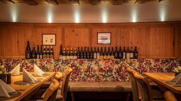 Ein gemütliches Restaurant mit Holzwänden und bunten Blumenmustern. Auf einem Regal sind viele Flaschen angeordnet, und die Tische sind geschmackvoll gedeckt.