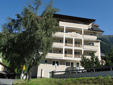 A modern building with several floors and balconies. Surrounded by trees and a mountainous landscape in the background.