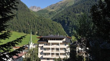 A picturesque mountain landscape with green forests and impressive peaks. In the foreground stands a modern building next to a small church.