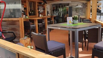 A cozy outdoor area of a restaurant with wooden decor and modern furniture. Colorful flowers and a sunshade awning create an inviting atmosphere.