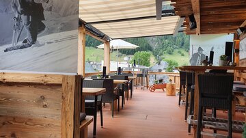 A cozy restaurant with wooden decor and a view of green mountains. The terrace is inviting and offers space for guests.