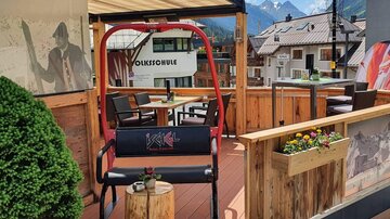 A cozy terrace with a red rocking chair and beautiful wood paneling. In the background, the mountains and a clear blue sky can be seen.