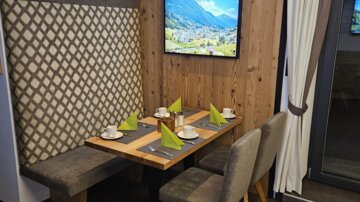 A cozy dining area with wooden details and a modern seating arrangement. Green napkins and cups are placed on the table, while in the background a television featuring a landscape picture is visible.