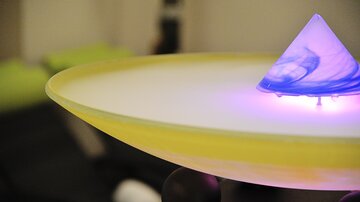 A modern table lamp with a glowing pyramid and a round, yellow base. The pyramid displays soft colors and creates a relaxed atmosphere.